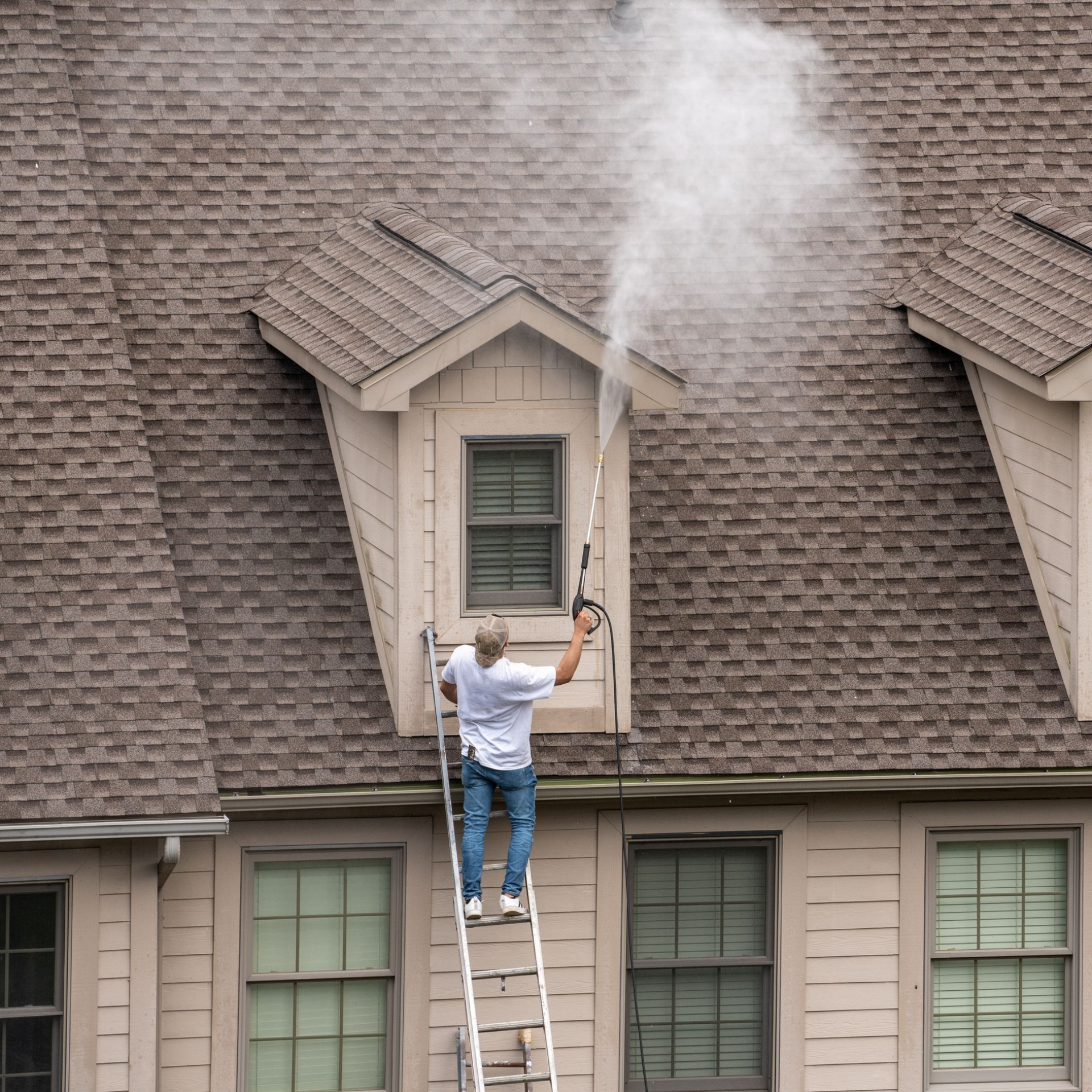 Man on a ladder pressure washing a roof, spraying water vapor. Beige house with brown shingles. Man on a ladder pressure washing a roof, spraying water vapor. Beige house with brown shingles.