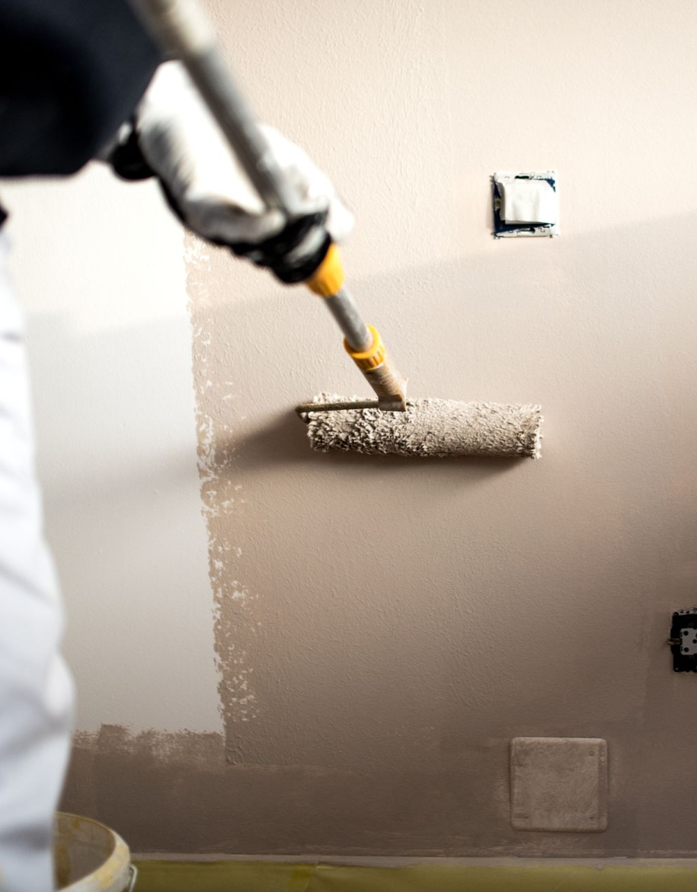 Person painting a wall with a roller, wearing gloves and white pants, in a room with electrical outlets.