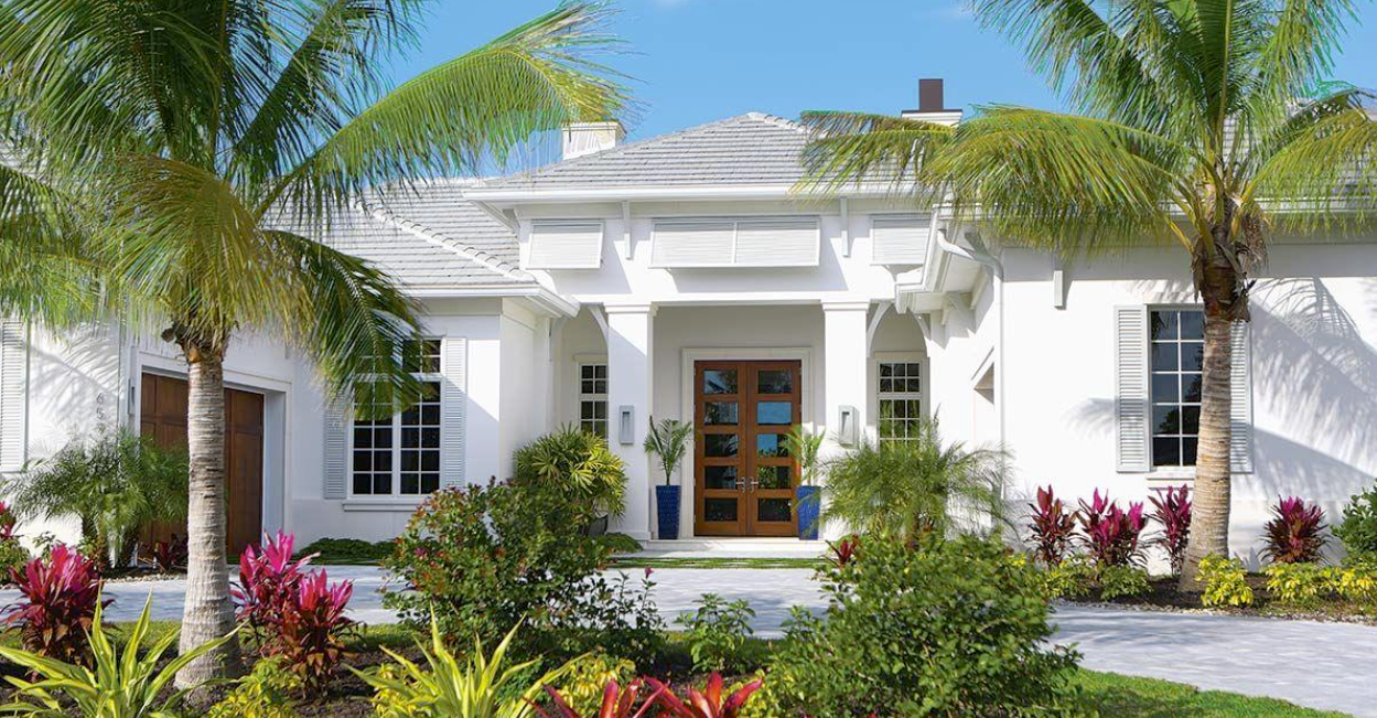 White house with wooden door, palm trees, and colorful plants under a blue sky.