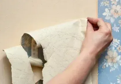 Hand peeling off beige wallpaper from a wall, revealing a floral blue wallpaper underneath.