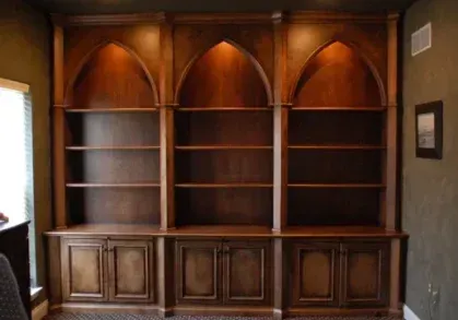 Built-in wooden bookcase with arched shelves, cabinets below, and interior lighting against a dark wall.