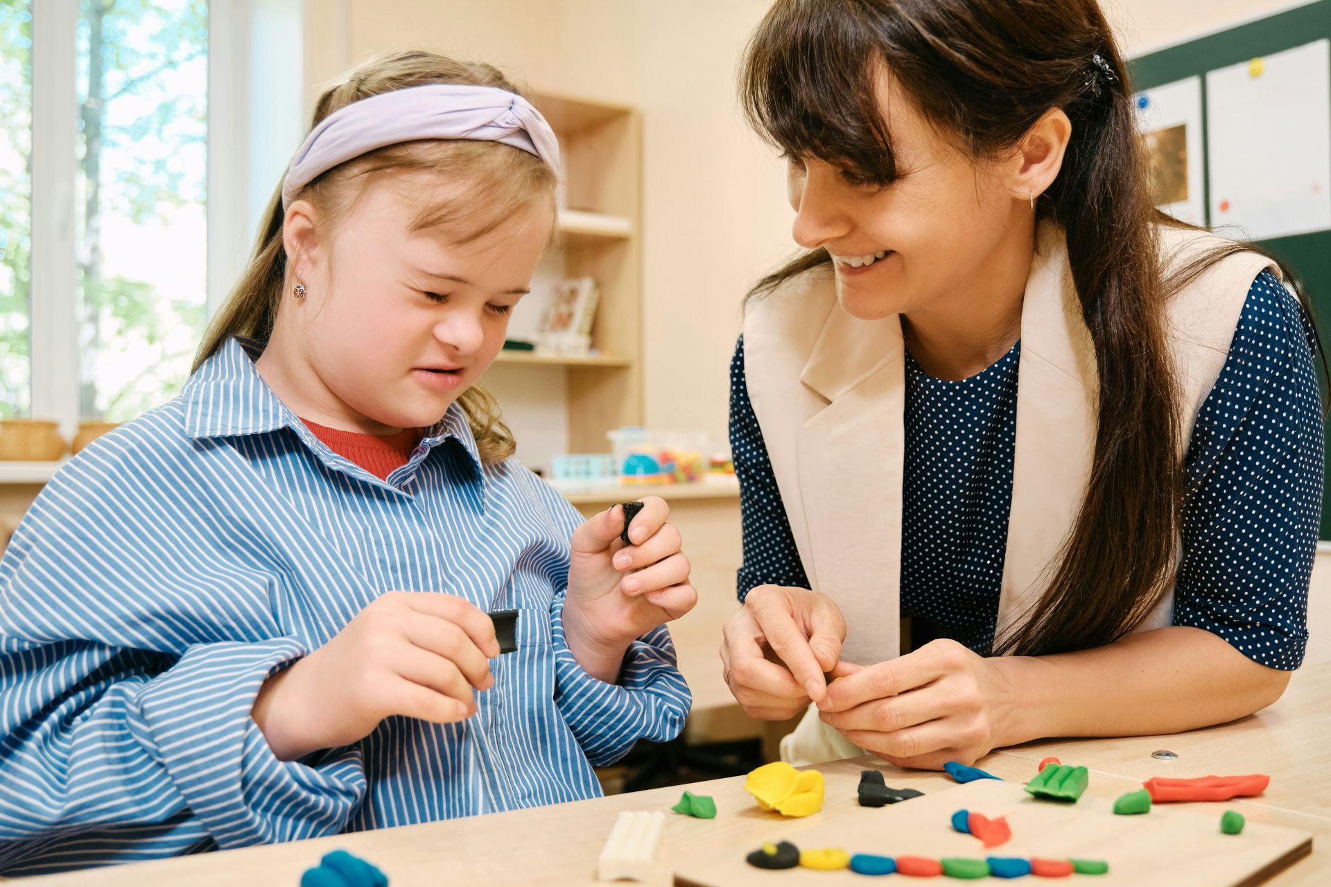 A student and teacher smile while working together with colorful modeling clay at a wooden desk in a classroom.