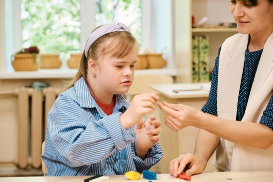 A teacher helps a student with Down syndrome shape colorful modeling clay during a focused, interactive classroom lesson.