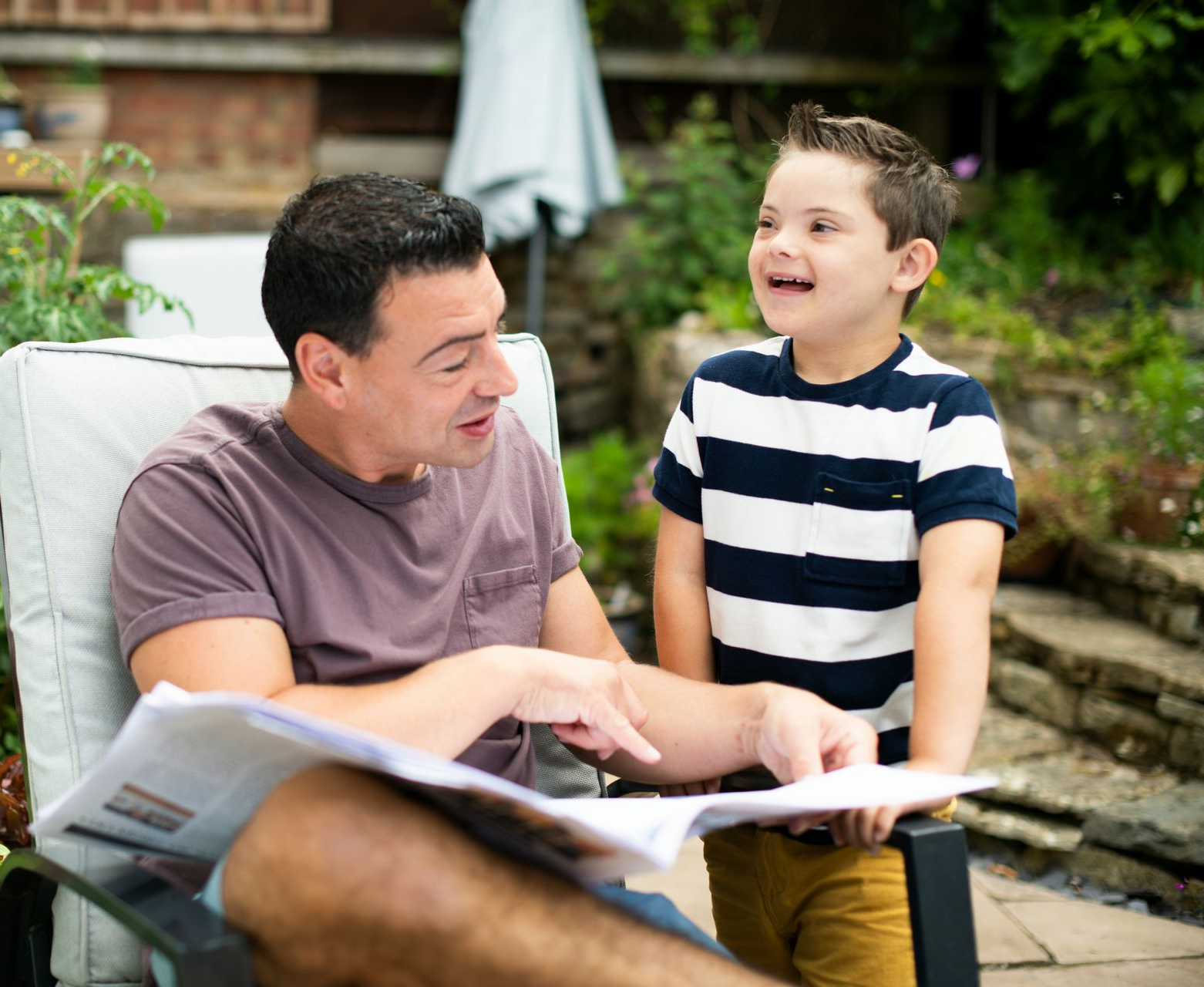 A man sitting in a garden chair points at a newspaper while talking to a young boy standing beside him.