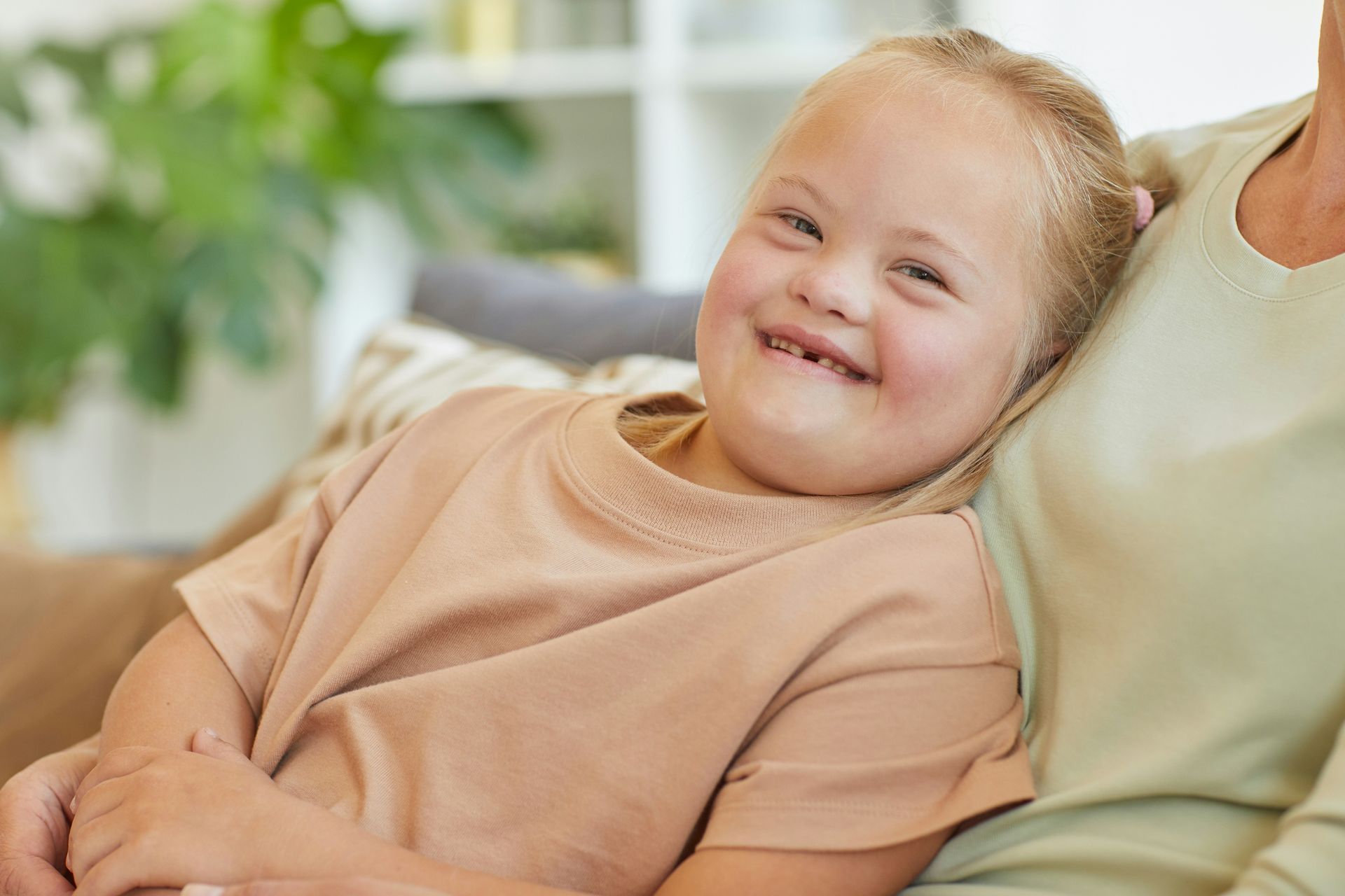 A smiling child with Down syndrome resting their head on an adult's shoulder while sitting together on a sofa.