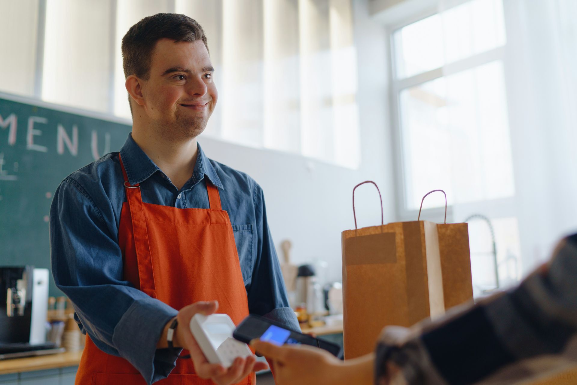 A smiling worker in an orange apron accepts a mobile phone payment from a customer next to a paper take-out bag.