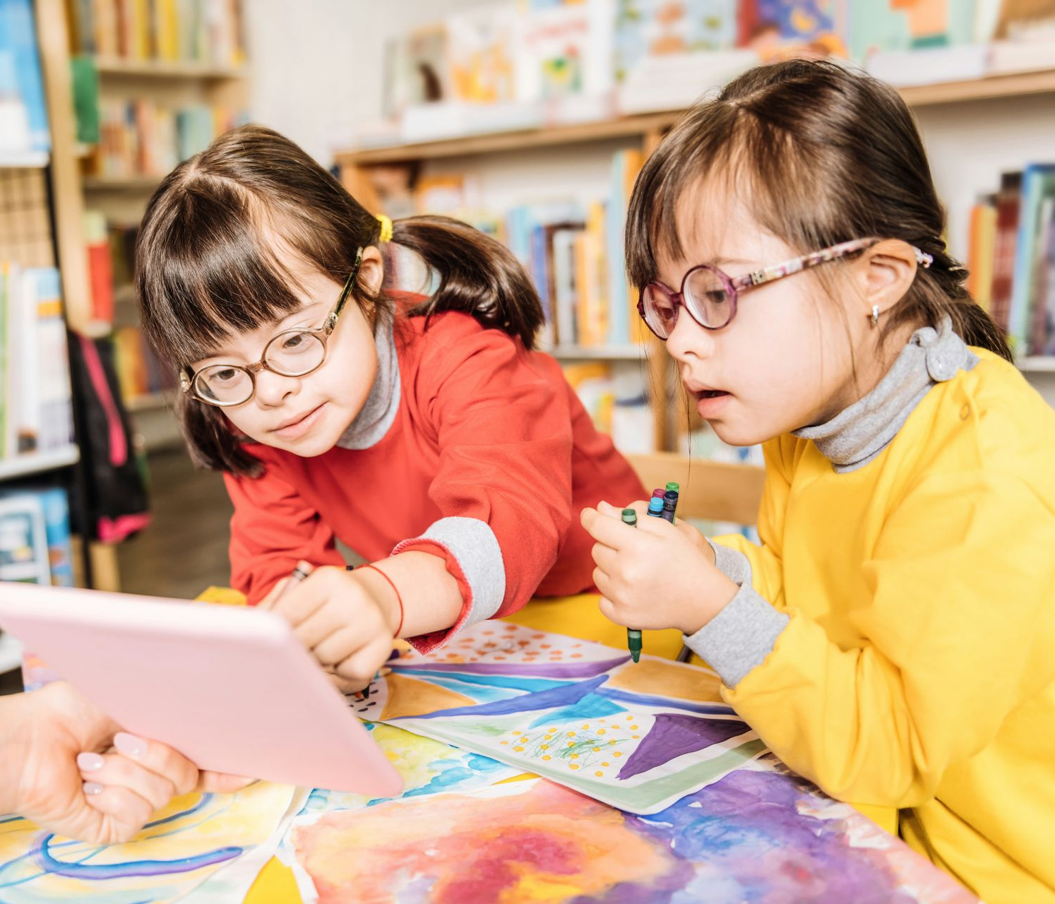 Two children wearing glasses and smocks interact with a tablet while working on a colorful art project in a library.