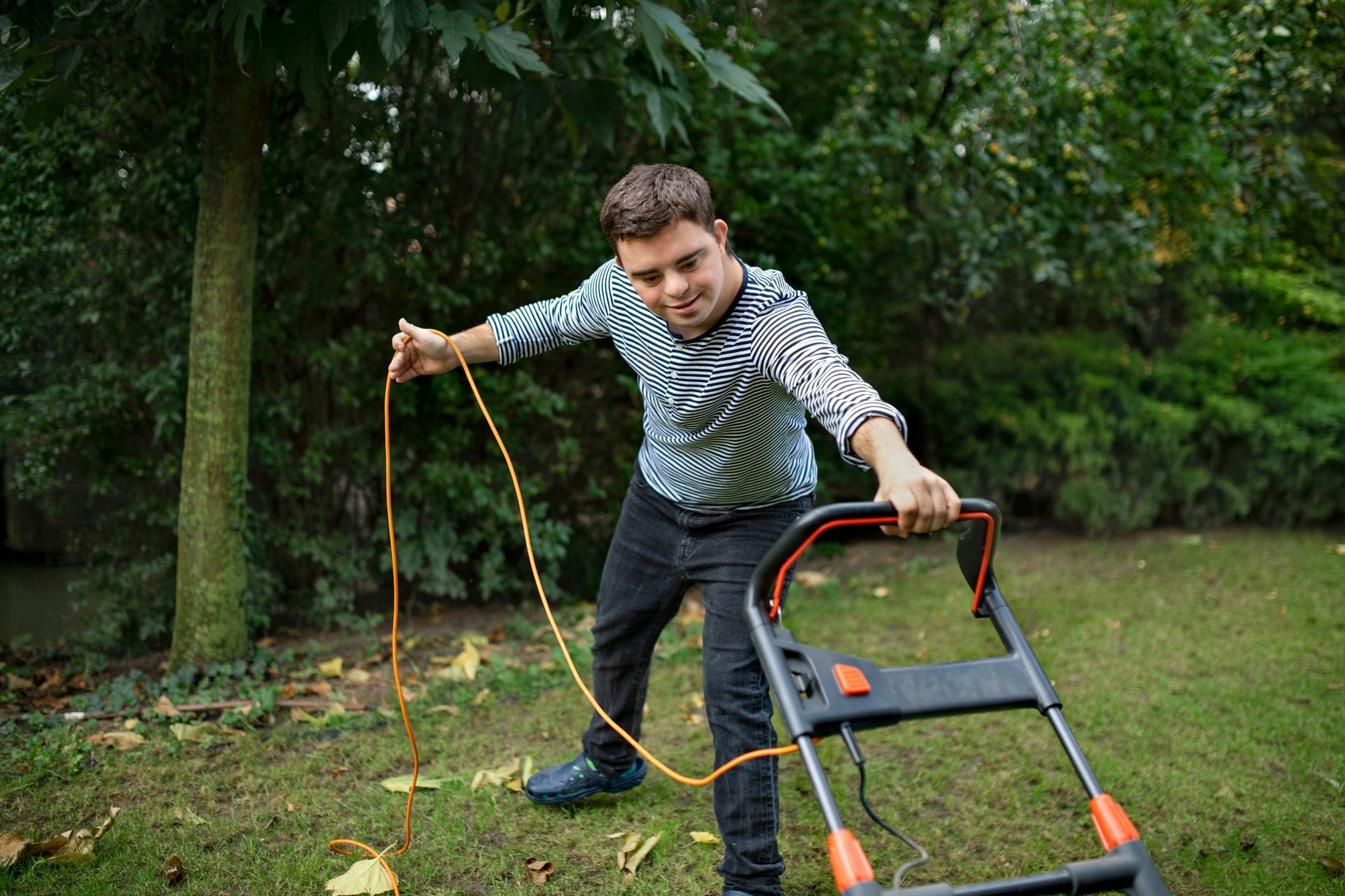 A person in a patterned long-sleeve shirt operates a corded lawn mower on a grassy lawn near trees.