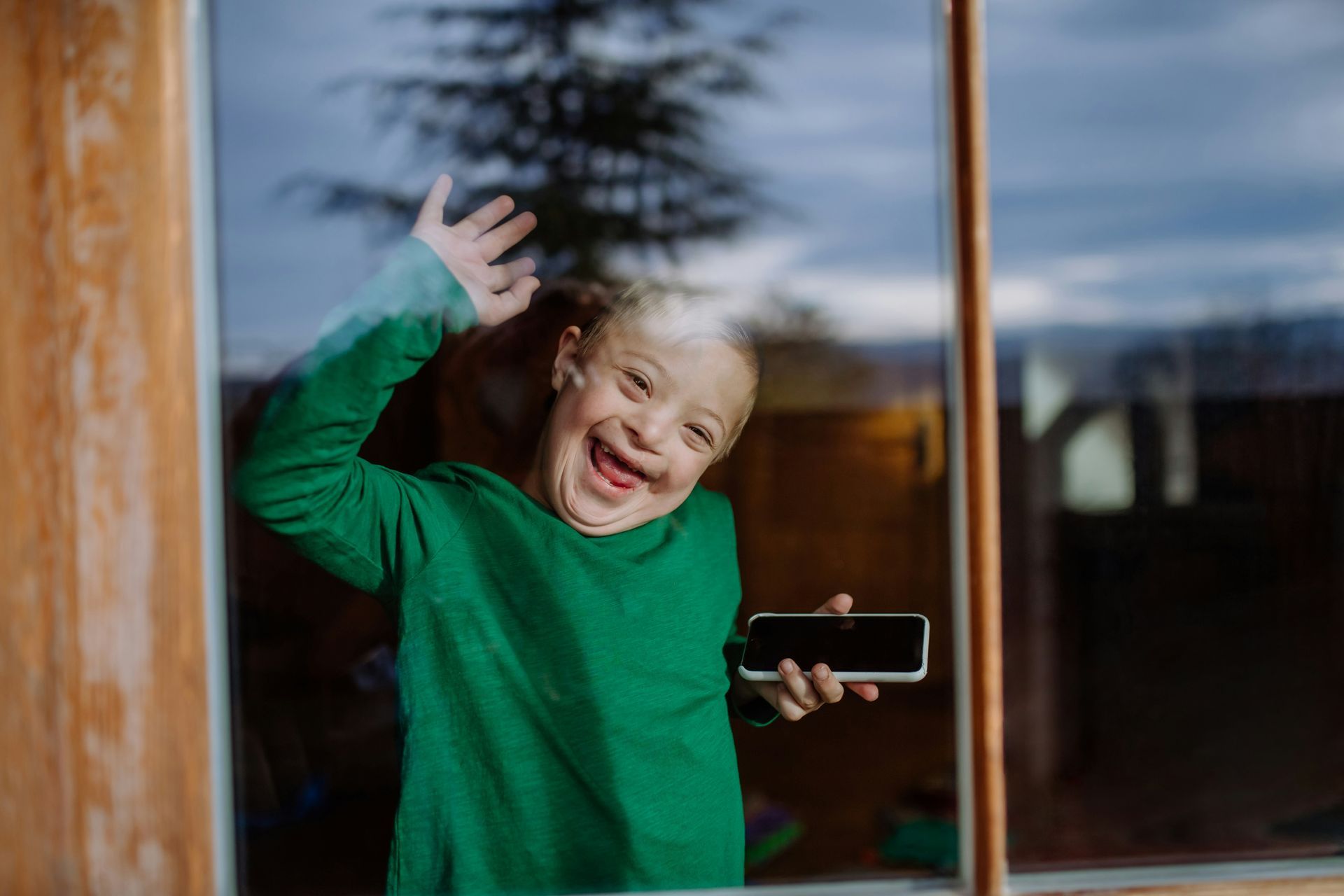 A person with a bright, joyful smile waves through a glass window while holding a smartphone.