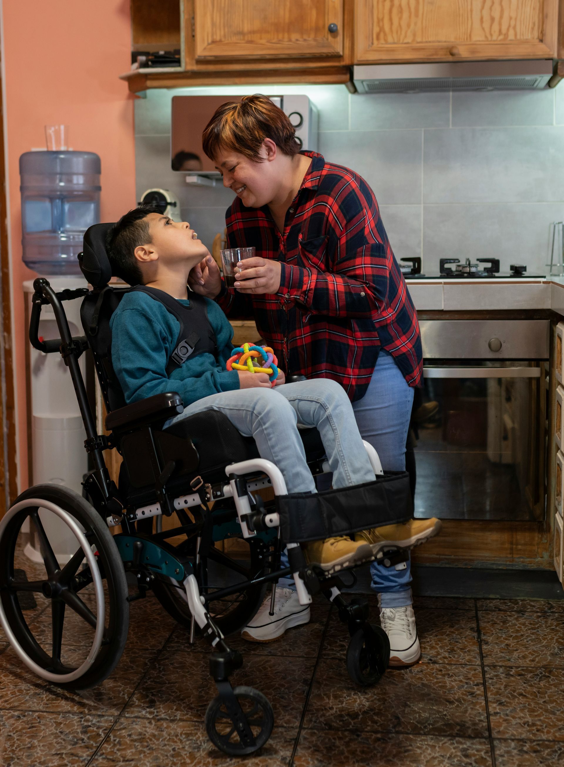 A person in a wheelchair smiles as another individual assists them by offering a drink in a kitchen.