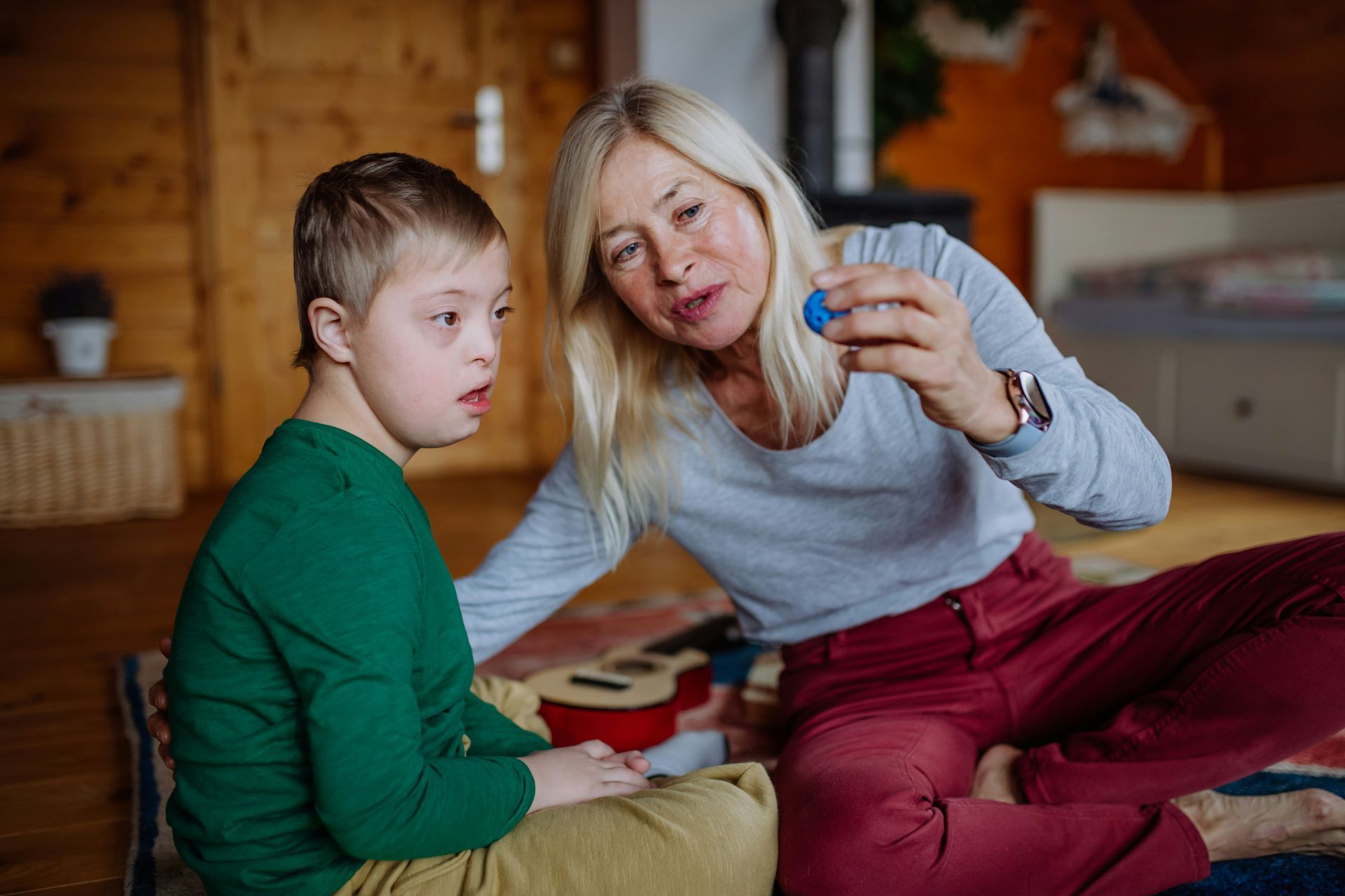 A woman holding a small blue object, interacting with a boy in a green sweater, both sitting on a floor in a room.