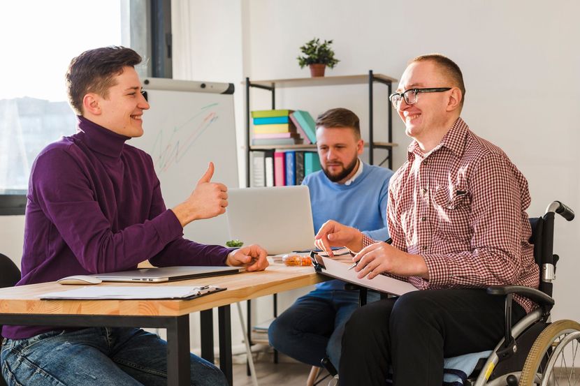 Three people in an office collaborate; one person in a wheelchair smiles as another gives a thumbs-up.