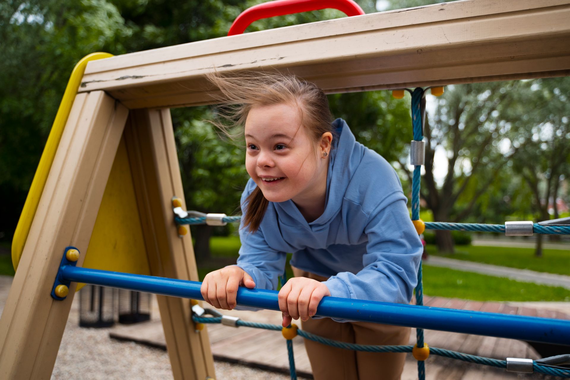 A young person with Down syndrome smiles while climbing a wooden jungle gym with a blue bar in a park.