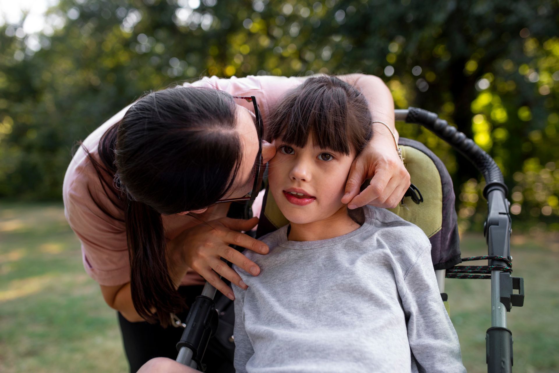 A person leans over to gently hold the face of a child sitting in a stroller in an outdoor, wooded setting.