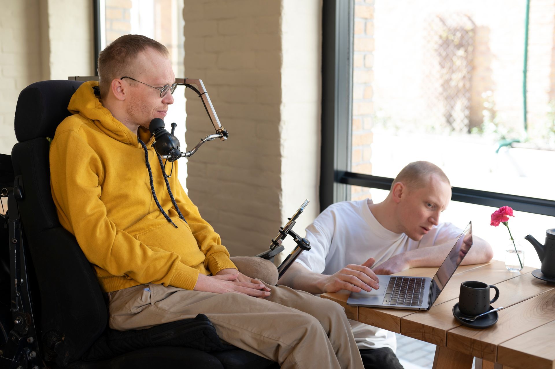 Two people seated at a table; one is in a wheelchair and the other types on a laptop next to a small flower and mug.