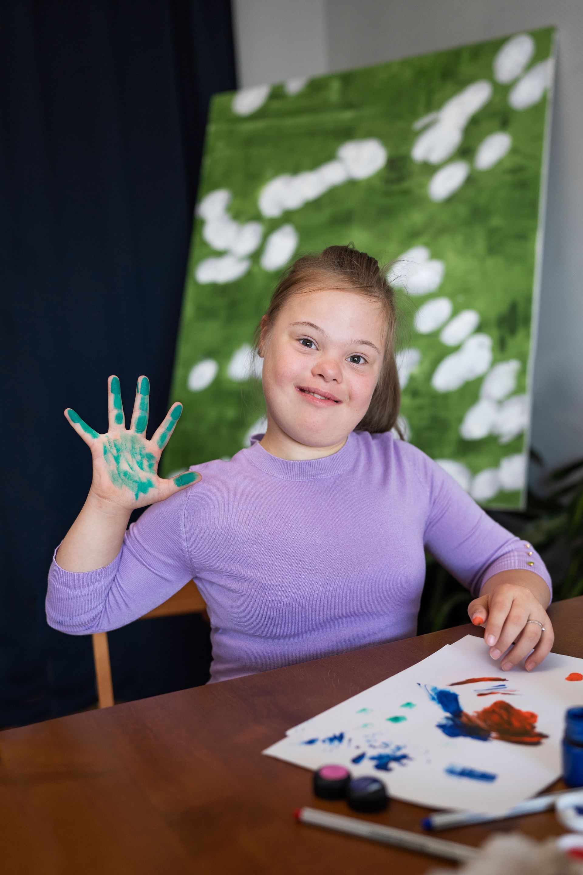 A person with a green-painted hand held up in a wave, smiling in front of a green canvas with white abstract shapes.