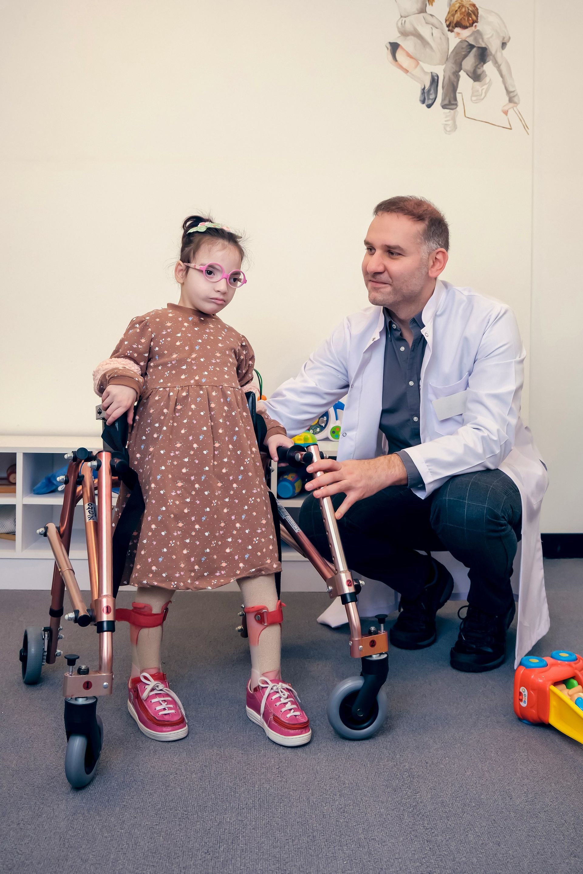 A clinician in a white lab coat assists a child using a copper-colored walker in a bright room with wall art.