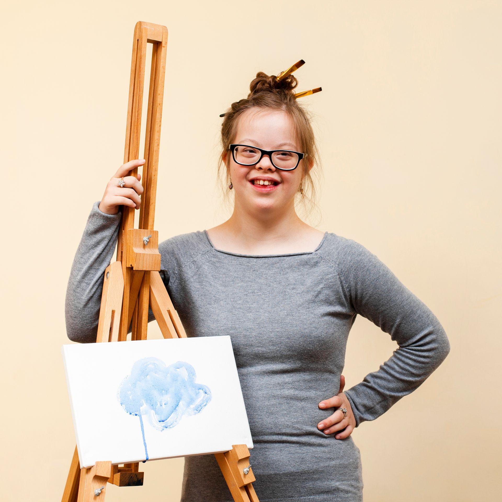 A person with glasses and a grey long-sleeved shirt stands smiling behind an easel with a painting of a blue shape.