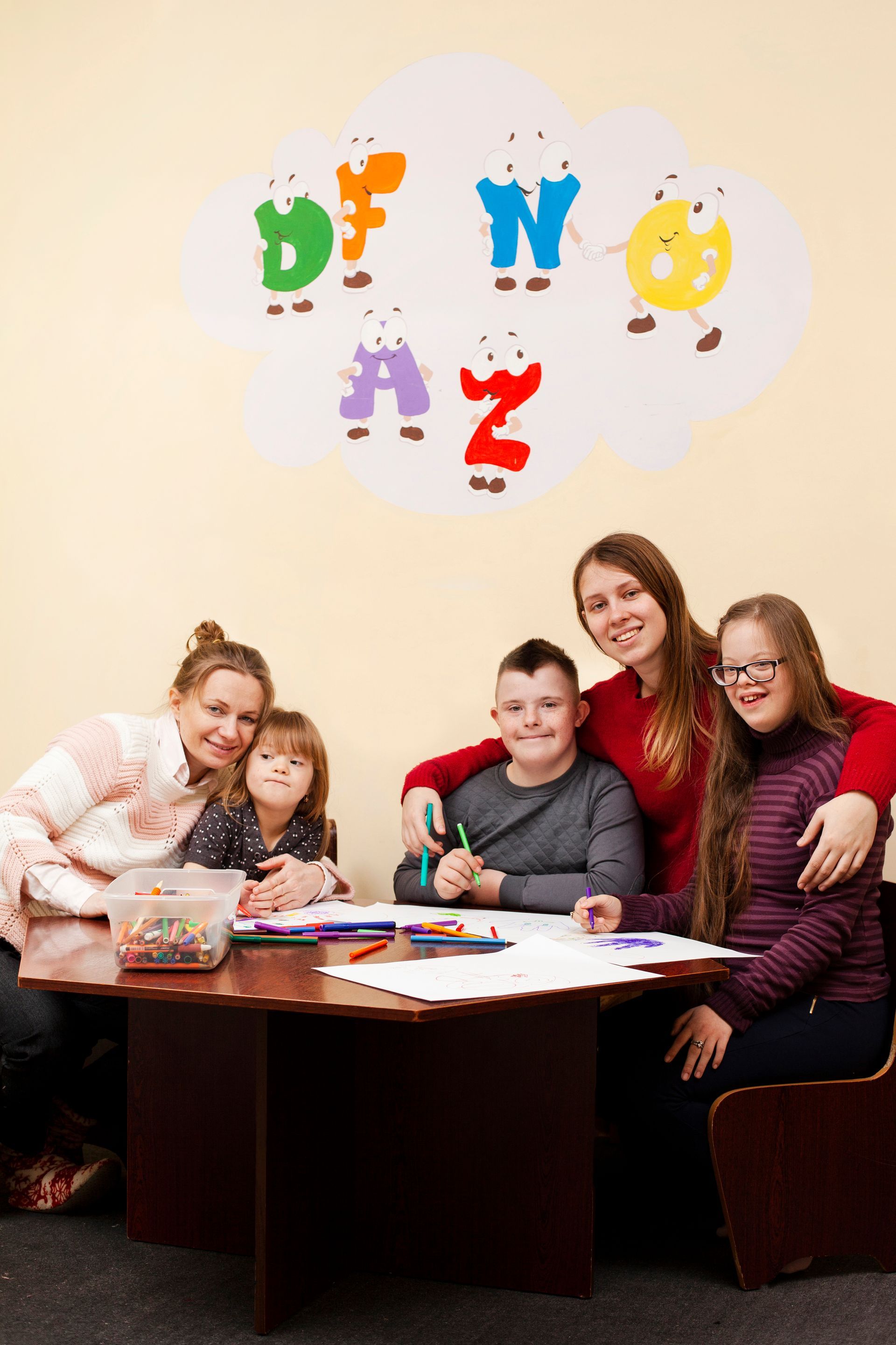 Two adults and three youth sit at a table working with craft supplies in a room with a decorative alphabet wall mural.