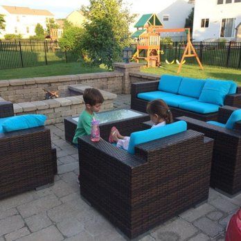Two children playing on outdoor furniture with blue cushions in a backyard with a playset and firepit.