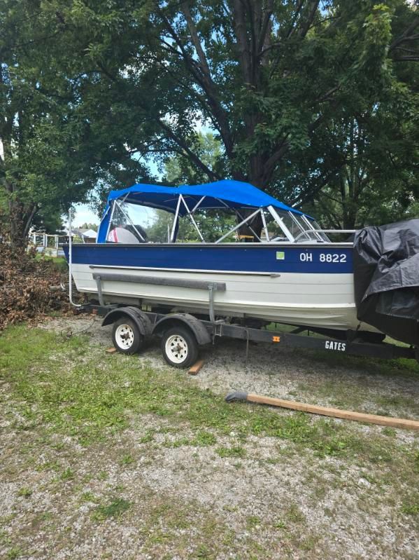 Blue and white boat on a trailer, under a blue canopy. The boat is in a grassy area with trees.