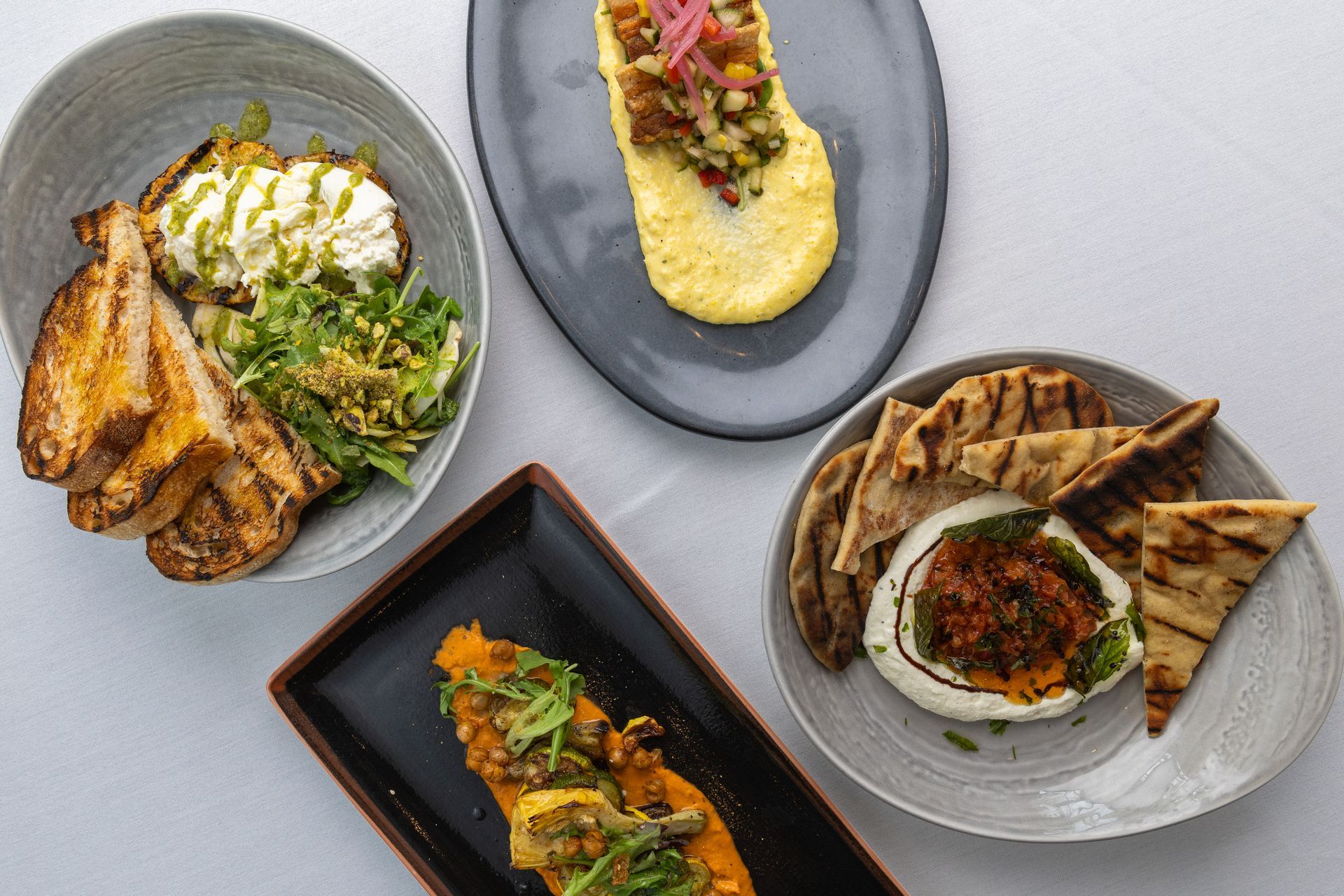 Four plates of food on a white surface. Dishes include toast, hummus, and grilled pita with cheese.