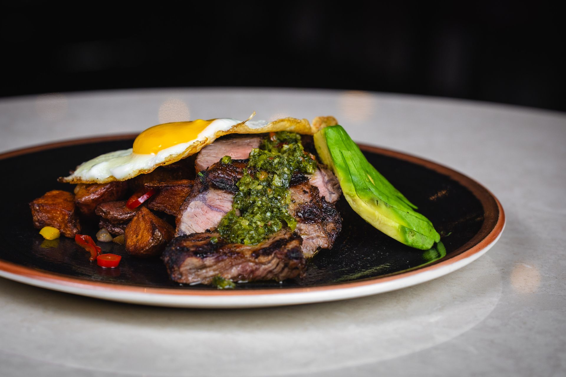 Steak with fried egg, avocado slice, and potatoes on a black plate, garnished with green sauce.