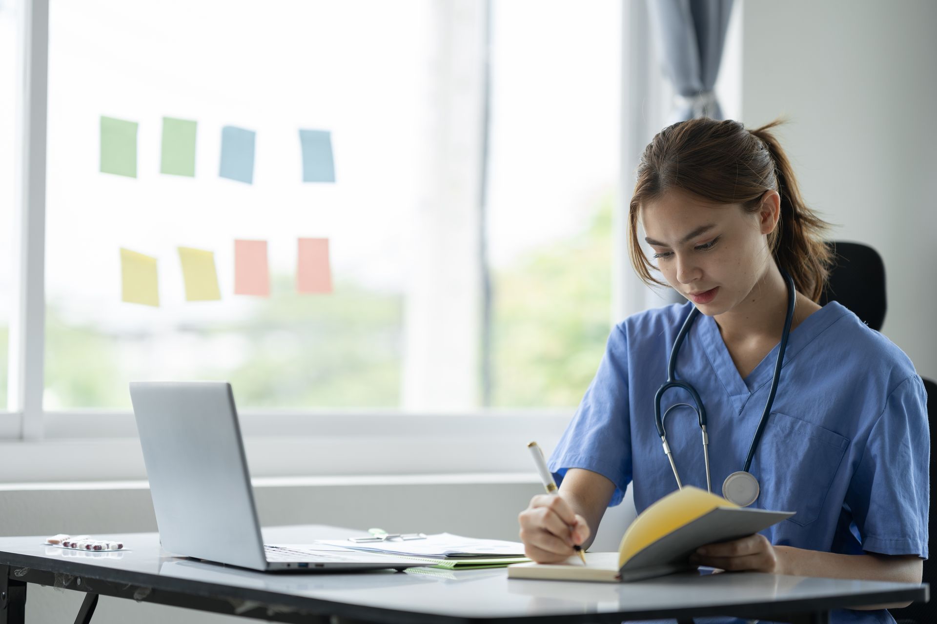 Kvinna i blå scrubs skriver vid ett skrivbord, öppen laptop, stetoskop, fönster med post-it-lappar.
