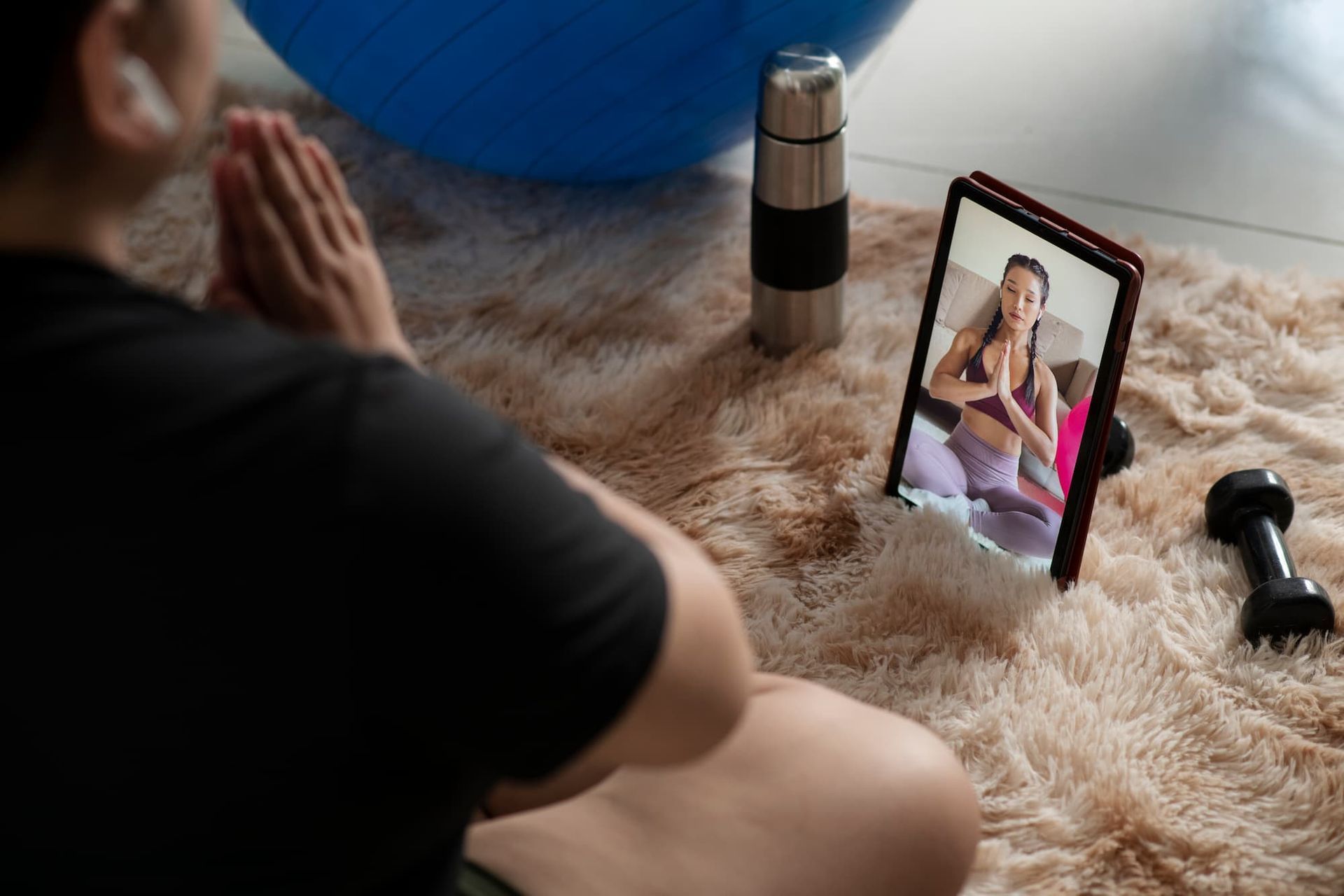 Person som gör yoga och följer onlineklass på surfplatta. Fluffig filt, träningsboll, vattenflaska och hantel syns.