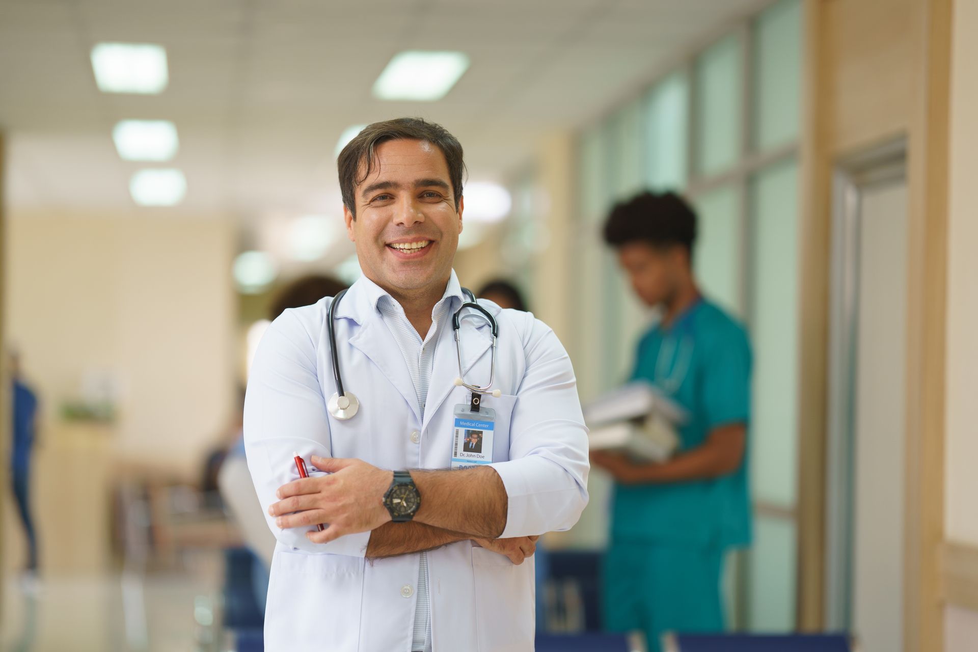 Läkare ler i sjukhuskorridoren, med armarna i kors, stetoskop och ID-bricka.