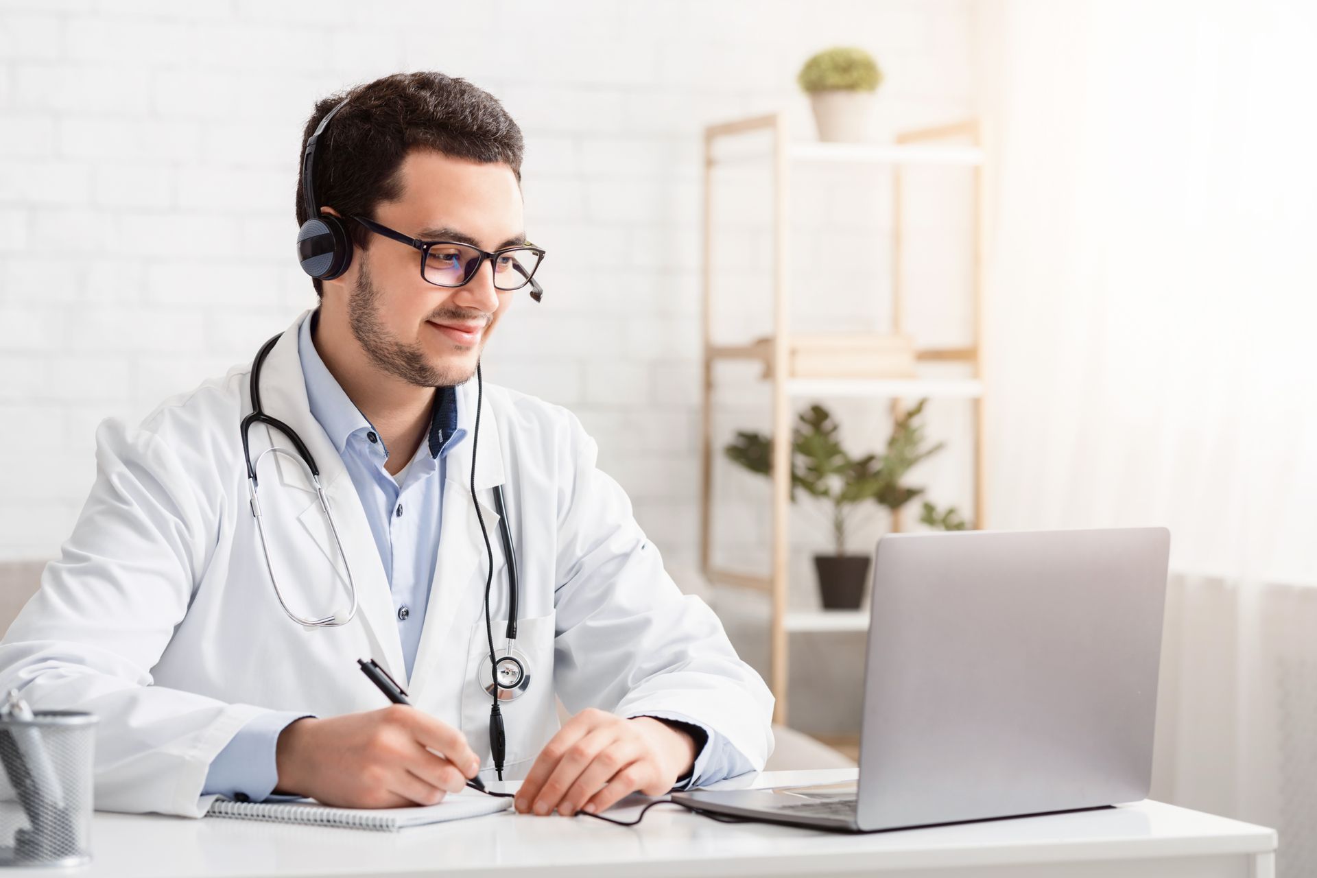 Läkare i vit rock, stetoskop, headset, antecknar under en telehealth-konsultation på en bärbar dator.