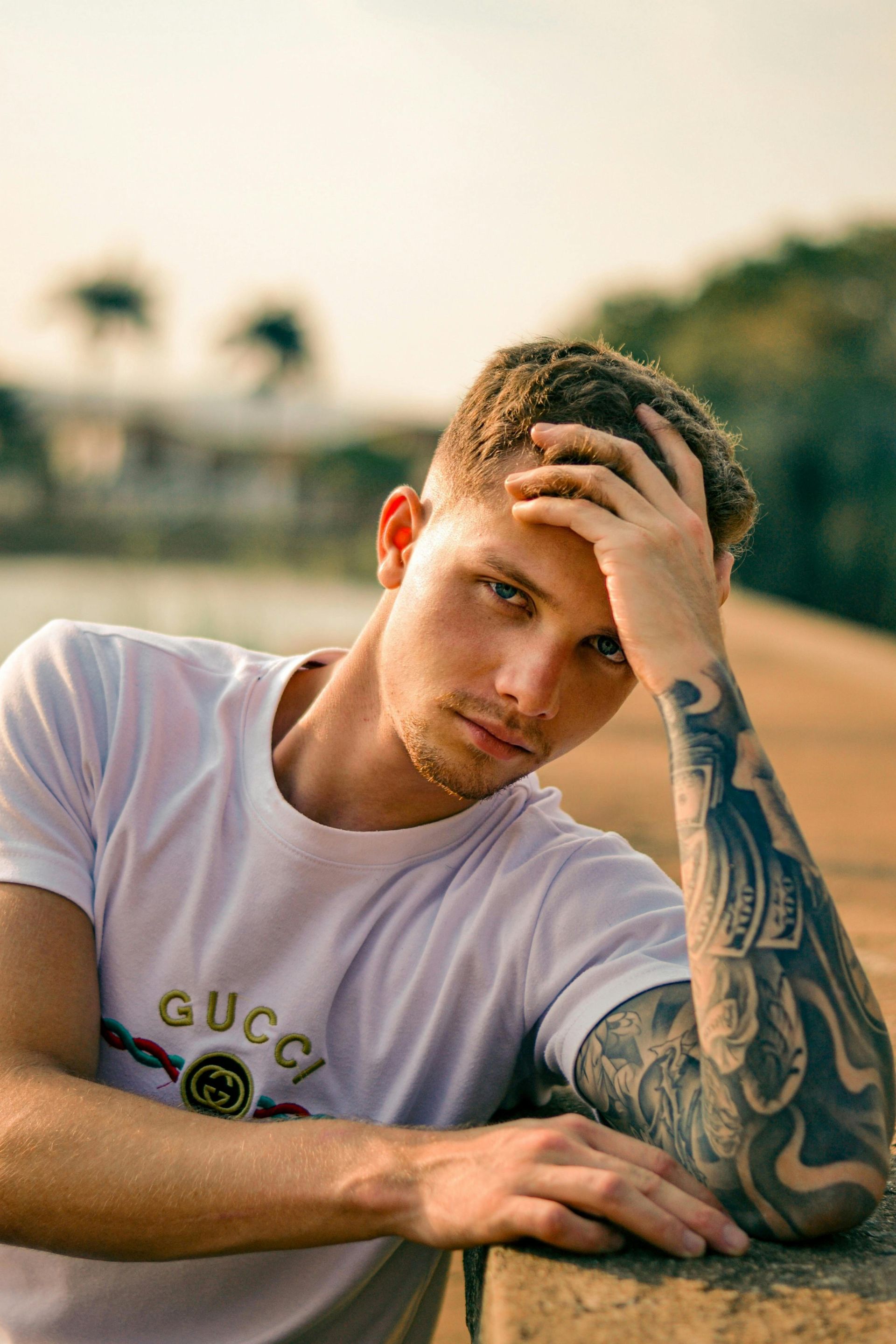 Man med armtatuering i vit Gucci-skjorta, handen på huvudet, tittar in i kameran. Utomhusmiljö.