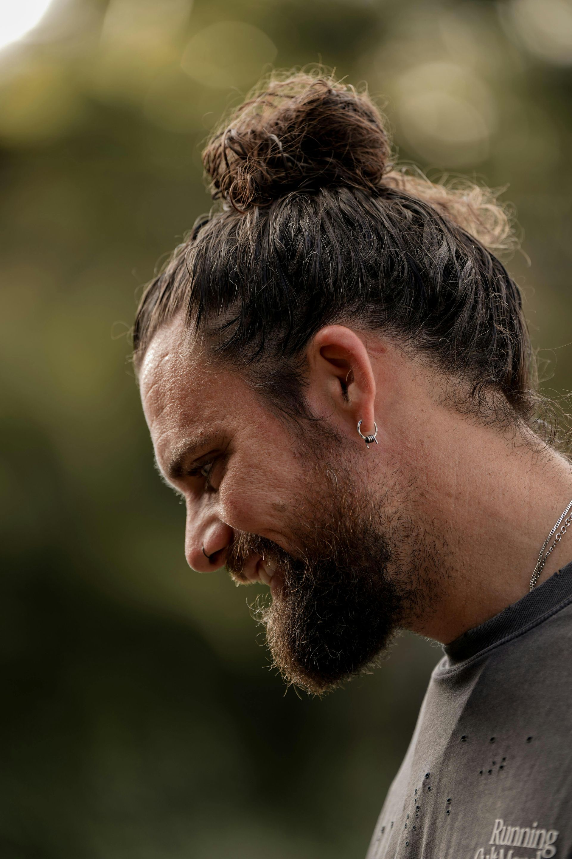 Man med mörkt hår i en knut, leende med skägg, utomhus.