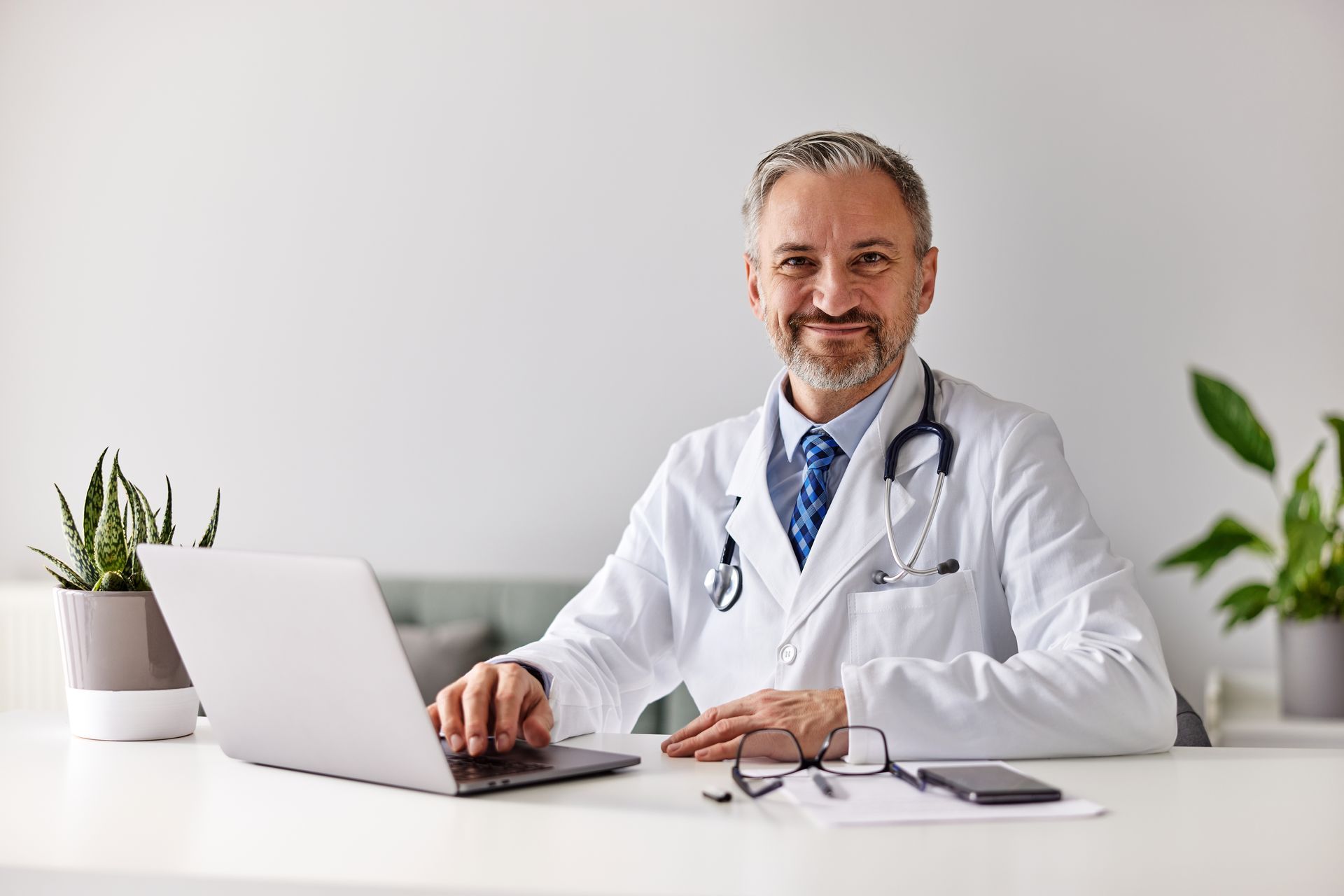 Läkare sitter vid skrivbord med laptop, stetoskop, leende och tittar in i kameran.