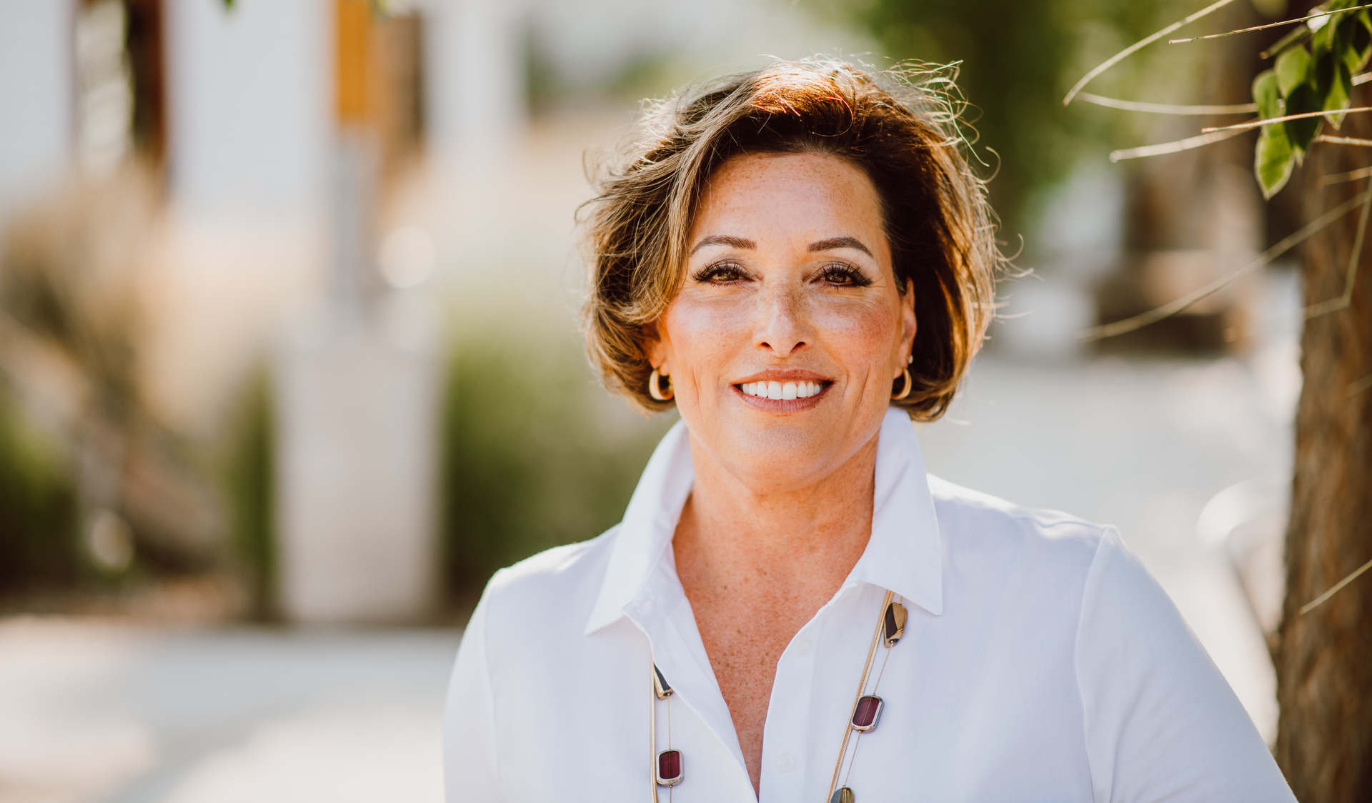 A woman in a white shirt is smiling for the camera while standing next to a tree.