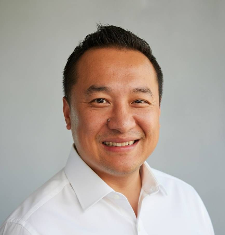Smiling man with dark hair, wearing a white shirt against a gray background.