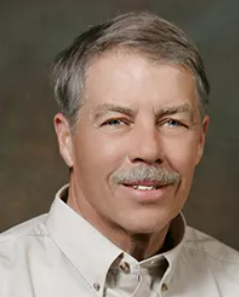 Man with short graying hair and mustache, wearing a light-colored collared shirt, smiling.