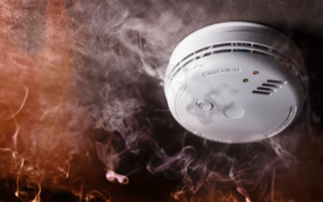 A smoke detector is surrounded by smoke and flames in a room.