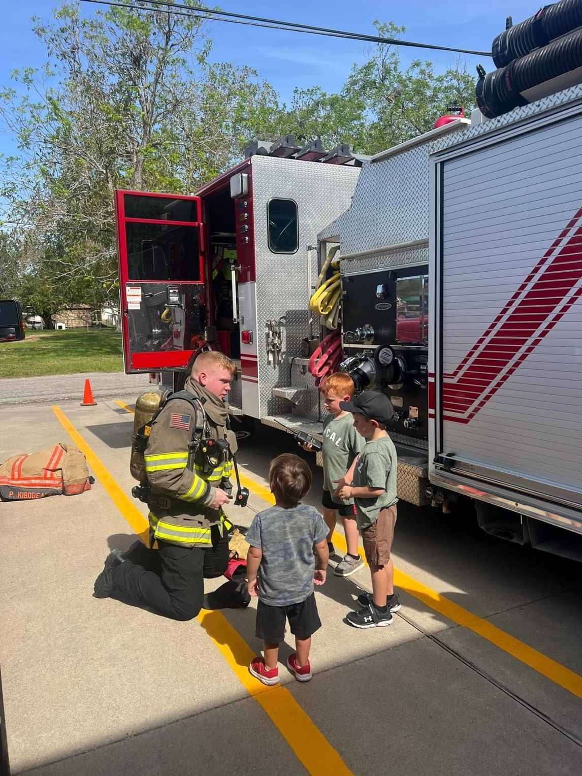 A firefighter is kneeling down to talk to a group of children in front of a fire truck.