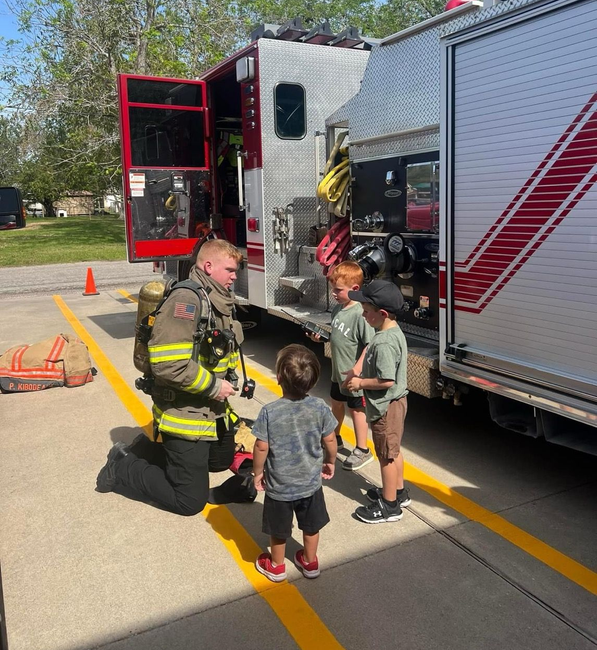 A firefighter is kneeling down to talk to a group of children in front of a fire truck.