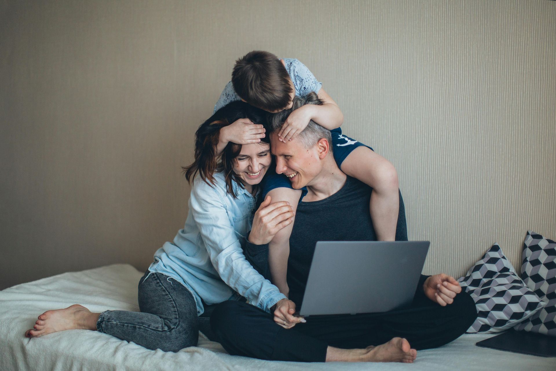 A family sits on a bed together, smiling while looking at a laptop computer.