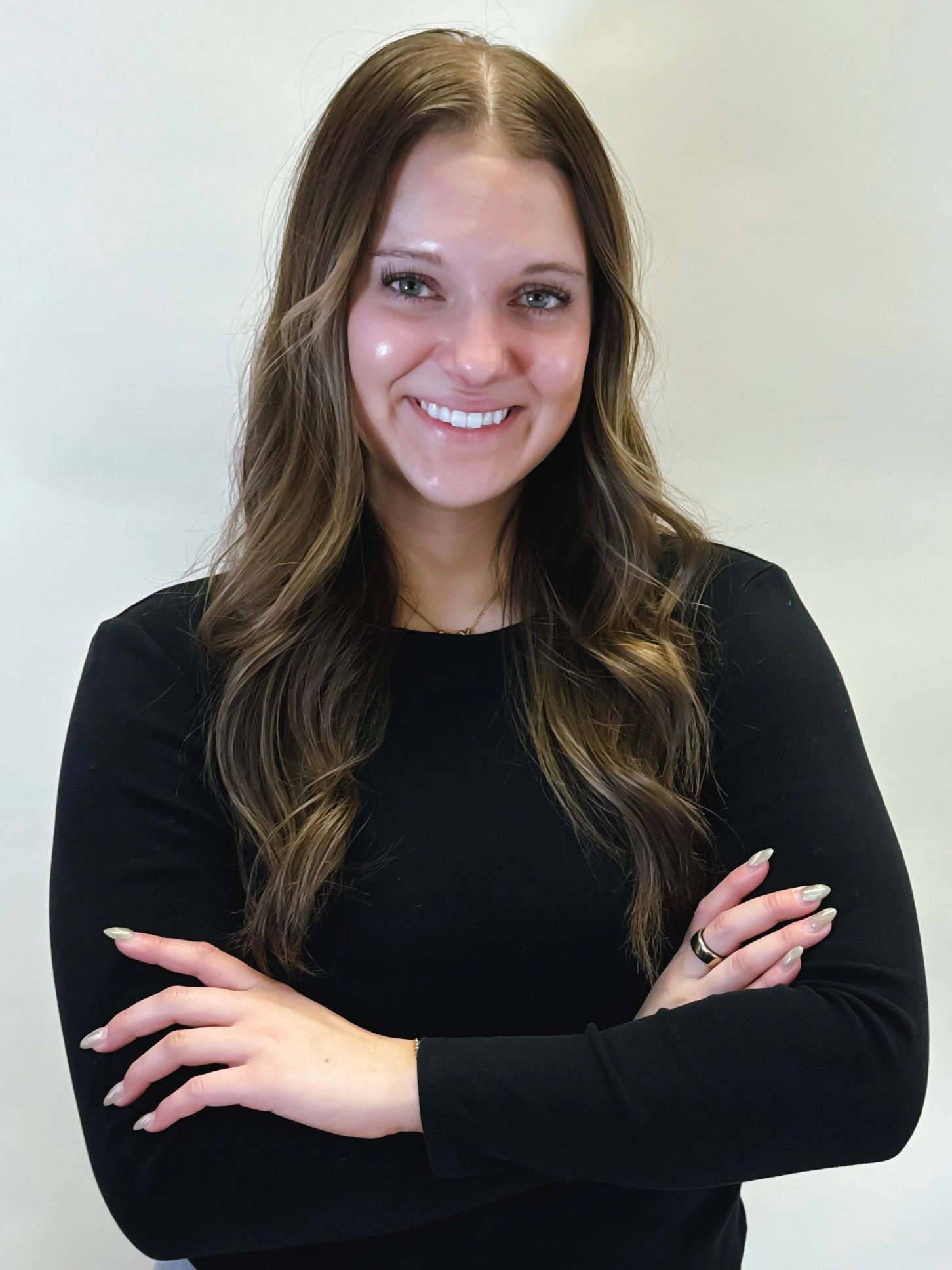 Smiling woman with long brown hair, arms crossed, wearing a black top against a light background