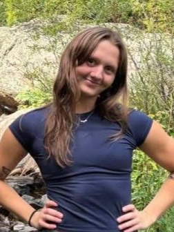 Woman in a navy shirt posing outdoors in front of greenery, hands on hips, smiling.