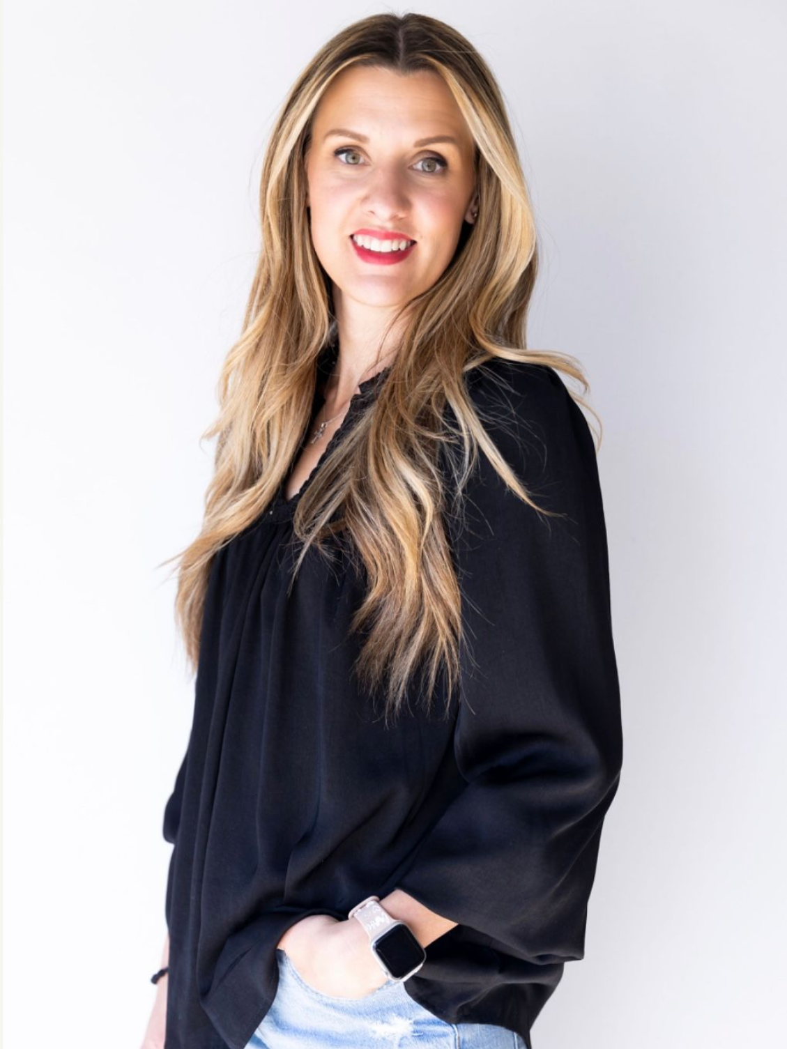 Woman in a black blouse and light jeans, standing against a white background and smiling softly