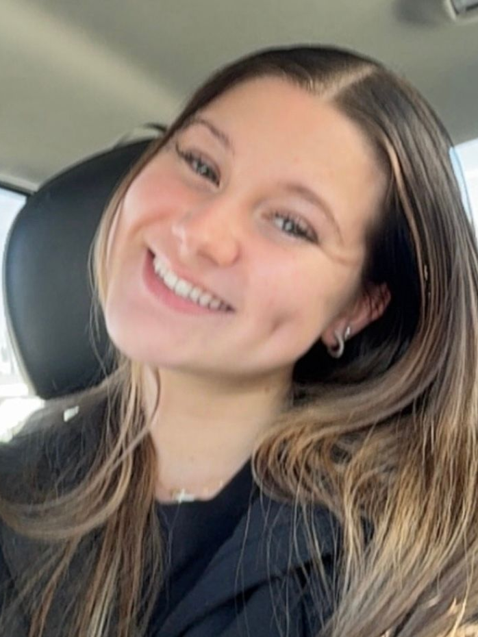 Smiling person with long brown hair sitting in a car, wearing a black top.
