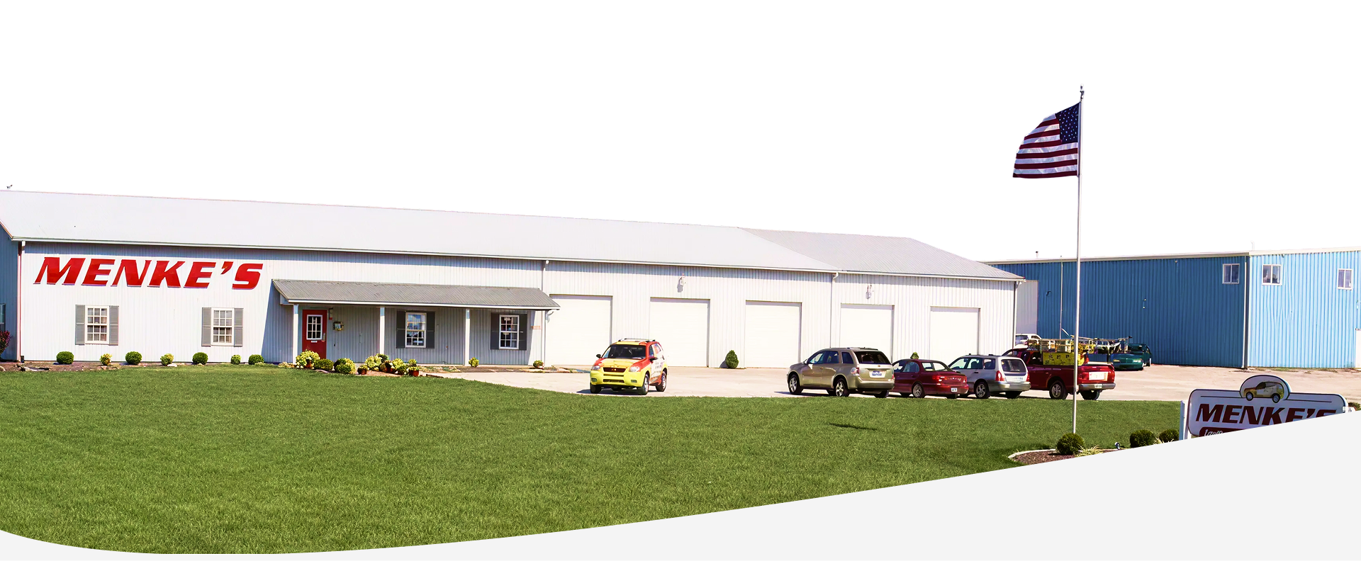Menke's building with a sign in front, cars, and American flag waving. | Menke's Automotive Repair