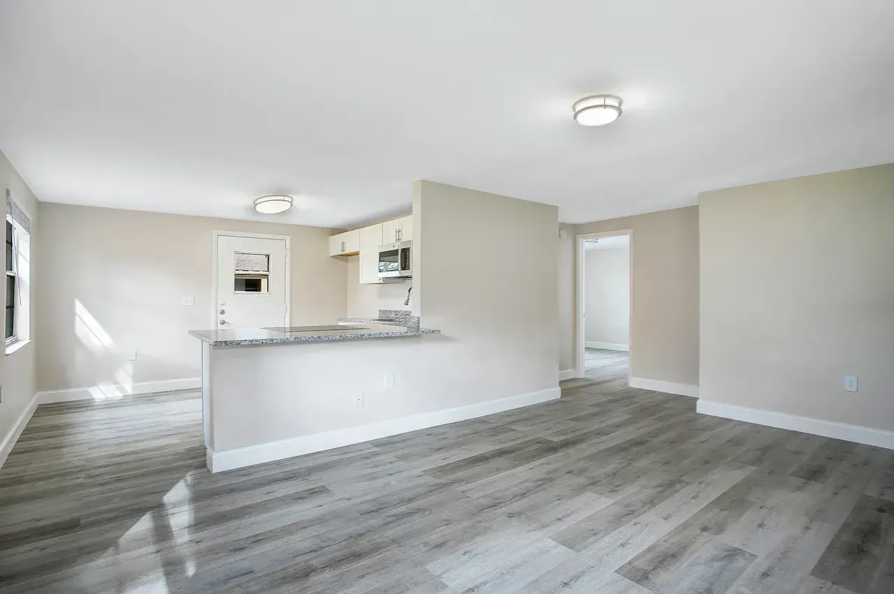 A living room with hardwood floors and a kitchen in the background.