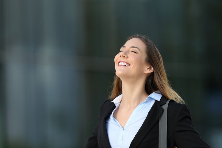 Woman in business attire smiles with eyes closed, outside a building.