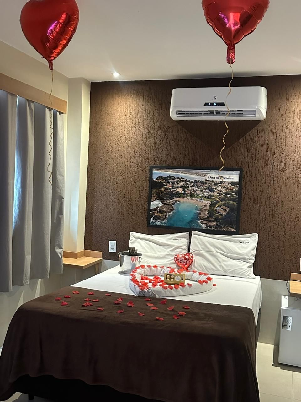 Quarto de hotel romântico com cama, balões vermelhos em forma de coração, pétalas e uma bandeja de sobremesas na cama.