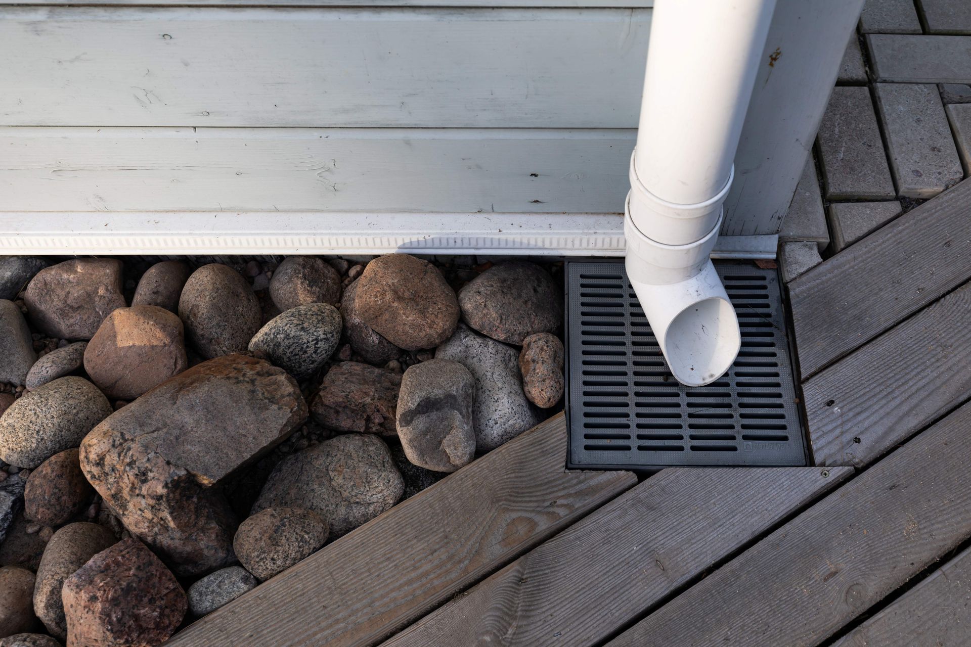 White drainpipe emptying into a grate on a wooden deck next to rocks.